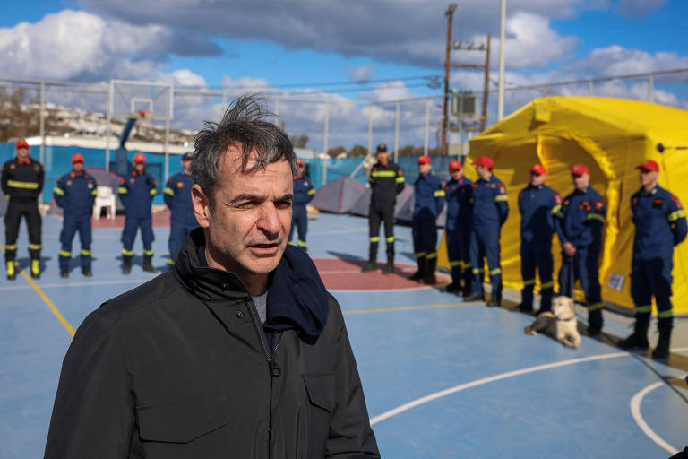 Um homem com cabelo bagunçado está em primeiro plano， falando ou se preparando para falar. Ao fundo， uma equipe de resgate， composta por homens e mulheres vestidos com uniformes azuis e bonés vermelhos， está alinhada. Eles estão em um campo de esportes， com uma tenda amarela visível ao lado. O céu está parcialmente nublado.