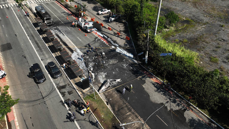 A imagem mostra uma vista aérea de uma estrada com um trecho danificado. Há veículos parados ao longo da via, incluindo caminhões e carros. Equipes de trabalho estão presentes, realizando reparos no asfalto, que apresenta marcas de queimadura. O ambiente ao redor é urbano, com vegetação ao fundo e sinalização de trânsito visível.