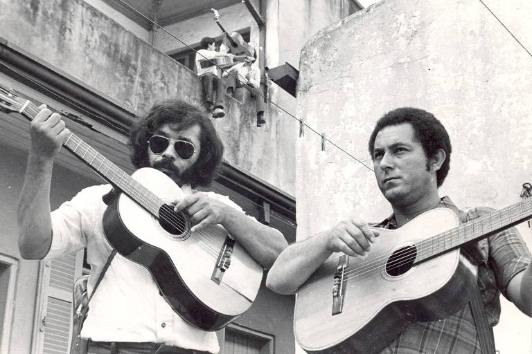 Dupla sertaneja Milionário e José Rico em cena do filme 'Na Estrada da Vida', em que o diretor Nelson Pereira dos Santos reconstrói a trajetória dos músicos. Na imagem, José Rico está com seus indefectíveis óculos escuros e segurando um violão. Milionário, por sua vez, também toca um violão e está vestido com uma camisa xadrez. Ao fundo, aboletados no alto de uma casa, dois jovens brincam com violões