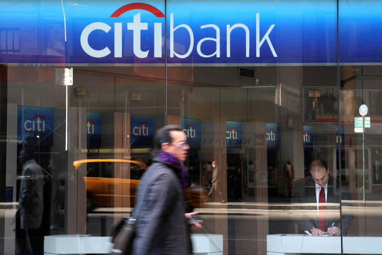 A imagem mostra a fachada de uma agência do Citi Bank. O logotipo do banco é visível na parte superior， com a palavra 039;citi039; em letras minúsculas e 039;bank039; em letras minúsculas， com uma linha vermelha acima da letra 039;i039;. Na frente da agência， um homem vestido com um casaco escuro e um cachecol roxo está caminhando. Ao fundo， um funcionário do banco está sentado em uma mesa.