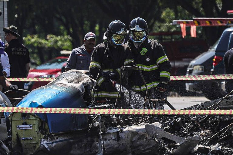 Imagem mostra dois bombeiros em meio aos destroços do avião， uma fita zebrada branca e vermelha isola a área