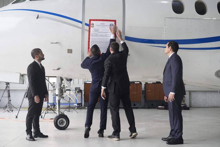 A imagem mostra um grupo de cinco pessoas em um hangar de aeronaves. Quatro delas estão vestidas com ternos escuros e observam enquanto uma pessoa， que está em pé em uma escada， coloca um documento em uma aeronave. A aeronave é de cor clara com detalhes em azul. Ao fundo， há algumas cadeiras empilhadas e parte do hangar visível.