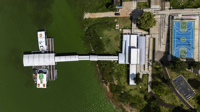 Imagem aérea de uma área recreativa à beira de um corpo d'água. À esquerda, há barcos atracados em um píer. À direita, uma quadra poliesportiva é visível, com linhas de marcação e cestas. O espaço ao redor é verde, com árvores e áreas de grama. Estruturas de edifícios estão presentes, conectadas por passarelas.