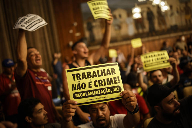 A imagem mostra um grupo de pessoas em uma manifestação， levantando cartazes. Um dos cartazes em destaque diz 039;TRABALHAR NÃO É CRIME!039; em letras grandes e amarelas. As pessoas parecem estar animadas e envolvidas na causa， com algumas segurando outros cartazes e gritando.