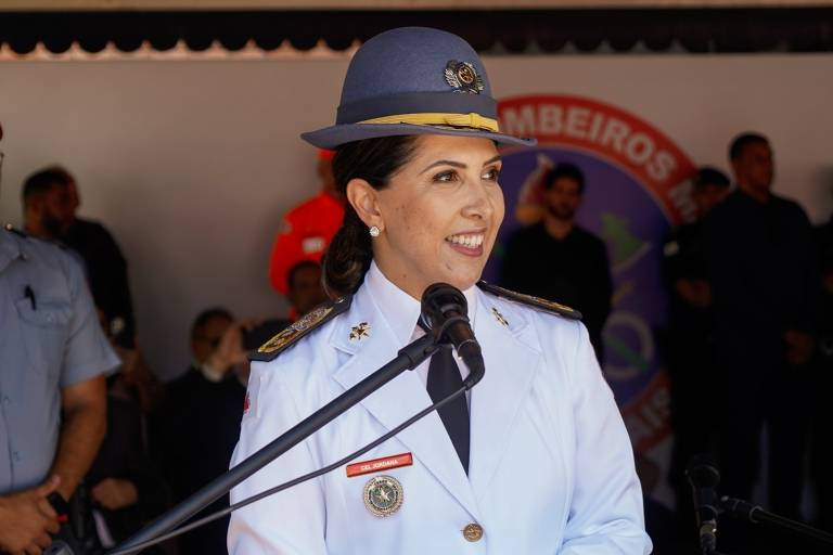 Uma mulher em uniforme branco， com um chapéu azul， está sorrindo enquanto fala ao microfone. Ela usa insígnias e um crachá que diz 039;CEL. JORDANA039;. Ao fundo， há pessoas em uniformes e um banner com a palavra 039;BOMBEIROS039;.