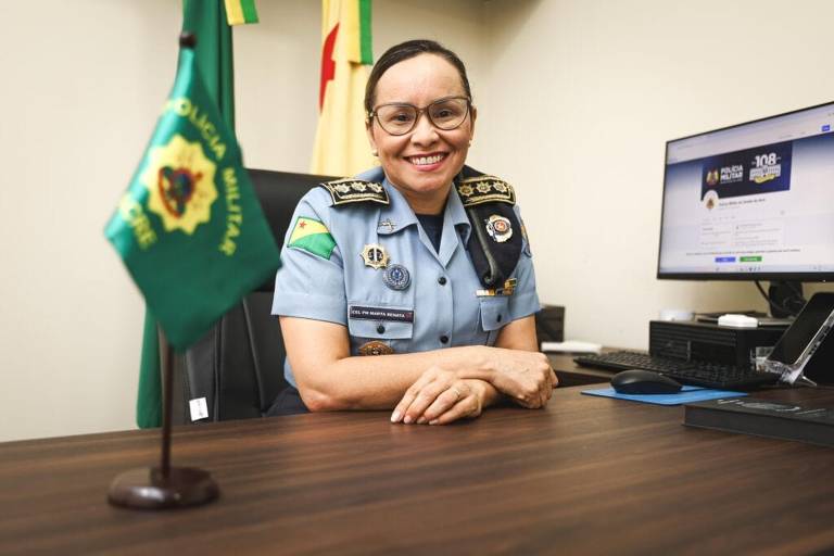 Uma oficial da Polícia Militar， vestindo um uniforme azul claro com insígnias， está sentada em uma mesa de escritório. Ela sorri para a câmera， com as mãos apoiadas na mesa. Ao fundo， há um computador com uma tela visível e bandeiras da polícia. A bandeira verde da Polícia Militar está em primeiro plano à esquerda.