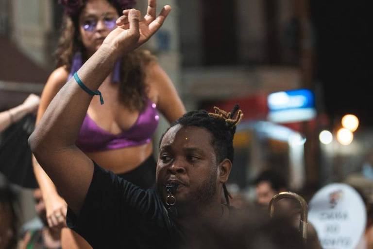 A imagem mostra um ambiente festivo à noite， com uma mulher em trajes de dança em cima de um homem. O homem está sorrindo e levantando a mão， enquanto a mulher parece estar se divertindo. Ao fundo， há várias pessoas e luzes coloridas， sugerindo uma celebração animada.