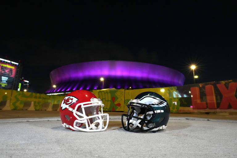 A imagem mostra dois capacetes de futebol americano， um vermelho com detalhes brancos e outro preto com detalhes verdes， posicionados lado a lado no chão. Ao fundo， um estádio iluminado em roxo está visível， com um céu noturno ao redor.