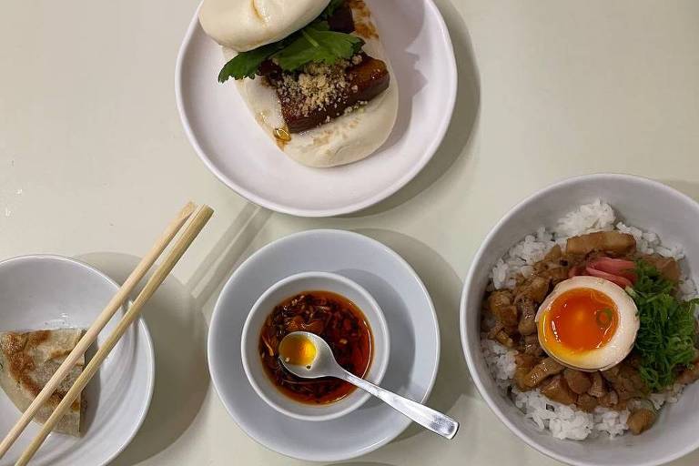 A imagem mostra uma mesa com pratos de comida. Em destaque, há dois baos em pratos brancos, um com carne e coentro, e outro com uma camada de carne frita. Ao lado, há um prato com arroz, carne, um ovo cozido com gema mole e vegetais. Também estão presentes um pequeno recipiente com molho e um par de hashis. Um pequeno prato com um bolinho frito está no canto inferior esquerdo.