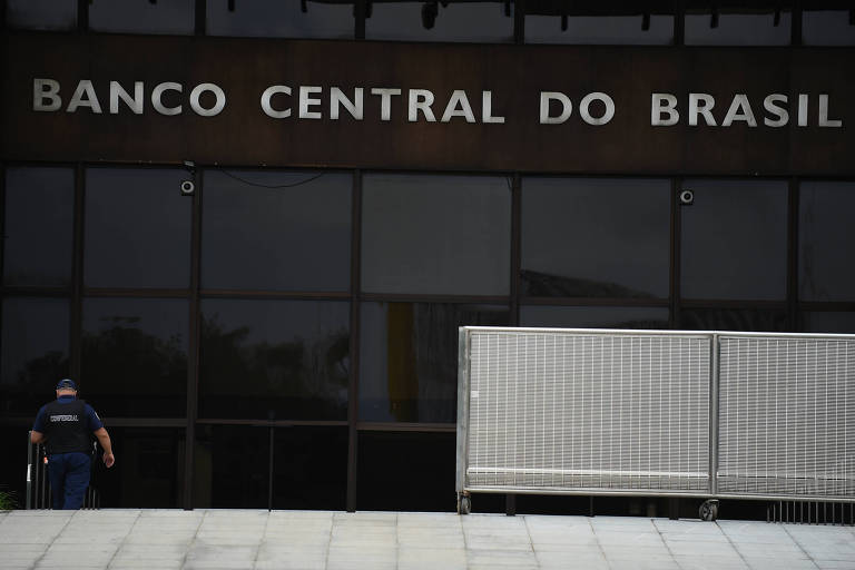 A imagem mostra a fachada do Banco Central do Brasil， com o nome 039;BANCO CENTRAL DO BRASIL039; em letras grandes e visíveis. Um homem， possivelmente um segurança， está se aproximando da entrada. Há uma barreira de segurança em frente à entrada， e as janelas refletem a luz do dia.