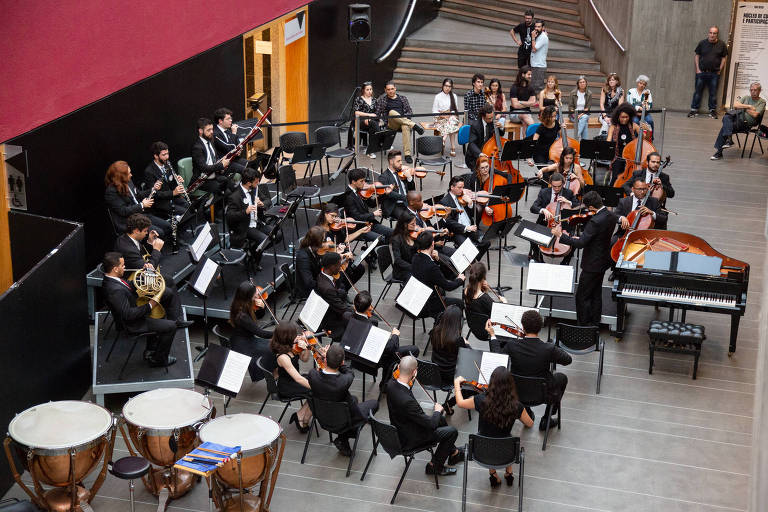 A imagem mostra uma orquestra se apresentando em um espaço amplo. Os músicos estão vestidos de preto e estão organizados em fileiras, com instrumentos como violinos, violoncelos, trompetes e um piano visível. Ao fundo, há escadas e algumas pessoas assistindo ao desempenho. O ambiente é moderno, com paredes de cores neutras e detalhes em vermelho.