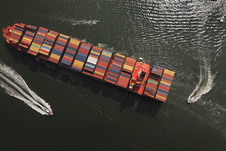 A imagem mostra um grande navio de carga laranja， coberto de contêineres coloridos， navegando em águas calmas. Ao redor， há pequenas embarcações brancas que também estão se movendo. A luz reflete na superfície da água， criando um efeito brilhante.
