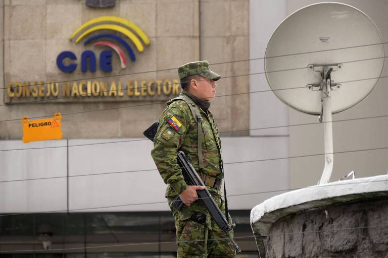 Um soldado vestido com uniforme militar camuflado está em pé， segurando um fuzil， em frente a um edifício que possui um logotipo colorido e a inscrição 039;CNE039; (Conselho Nacional Eleitoral). Ao fundo， há uma antena parabólica e uma parede de concreto.