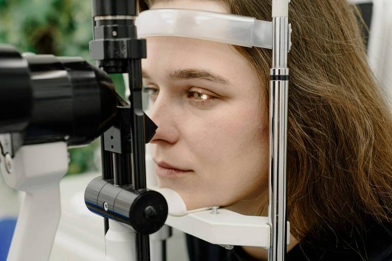 Uma jovem mulher está realizando um exame ocular em um consultório. Ela está posicionada em frente a um equipamento médico， com um suporte na cabeça e um dispositivo de exame próximo ao seu olho direito. O ambiente é clínico， com equipamentos de oftalmologia visíveis ao fundo.