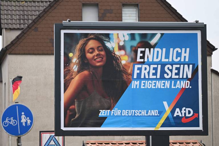 Um outdoor publicitário apresenta uma mulher sorridente com cabelo cacheado， em um fundo azul. O texto em destaque diz em alemão 039;Finalmente ser livre no próprio país039; e abaixo está escrito 039;Agora pela Alemanha039;. O logotipo do partido， AfD， também está presente. Ao fundo， há uma casa com telhado de duas águas e uma bandeira da Alemanha.