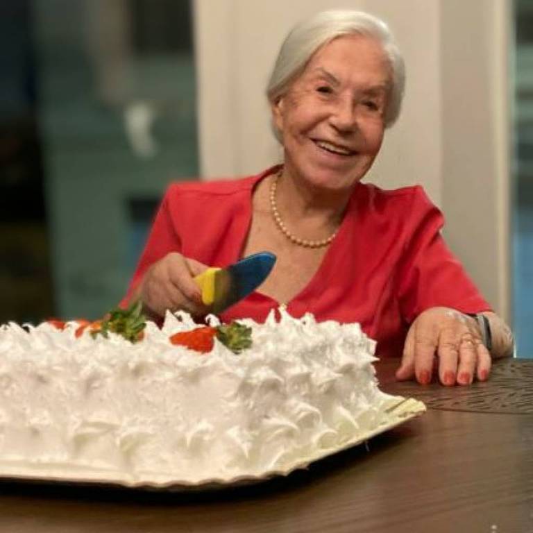 Uma mulher idosa com cabelo grisalho e curto está sorrindo enquanto se senta à mesa. Ela usa uma blusa vermelha e está segurando um bolo decorado com cobertura branca e enfeites coloridos. O ambiente é iluminado e há uma janela ao fundo.