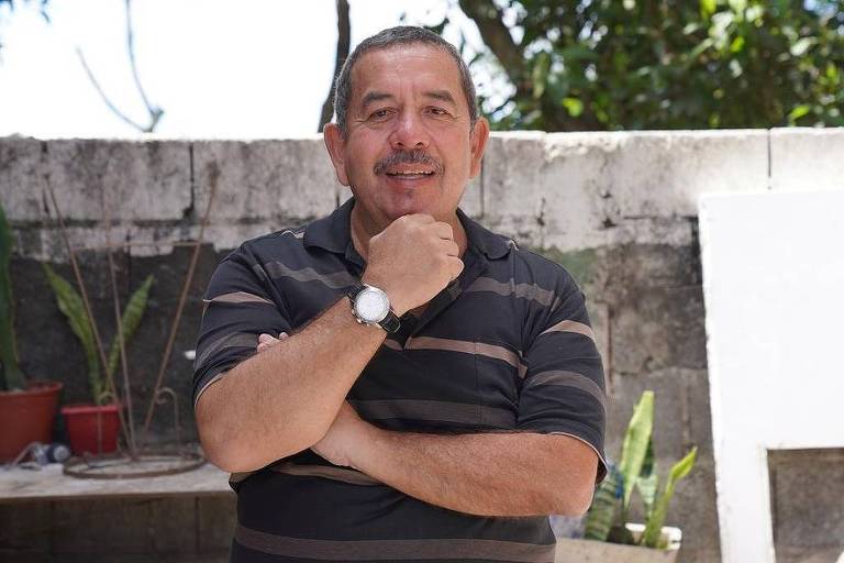 Um homem de meia-idade está posando para a foto com um sorriso. Ele está usando uma camisa listrada em preto e cinza e tem uma mão apoiada no queixo. Ao fundo， há uma parede de concreto e algumas plantas em vasos.