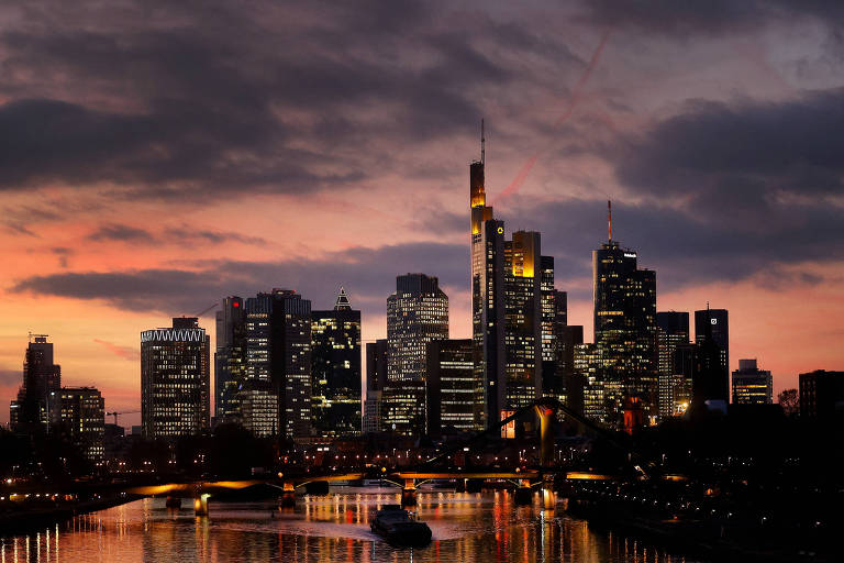 A imagem mostra um horizonte urbano durante o entardecer， com edifícios altos e modernos refletindo a luz do sol poente. O céu apresenta nuvens escuras e tons de laranja e roxo. Abaixo， há um corpo d039;água que reflete as luzes da cidade e os edifícios. A cena transmite uma atmosfera tranquila e urbana.
