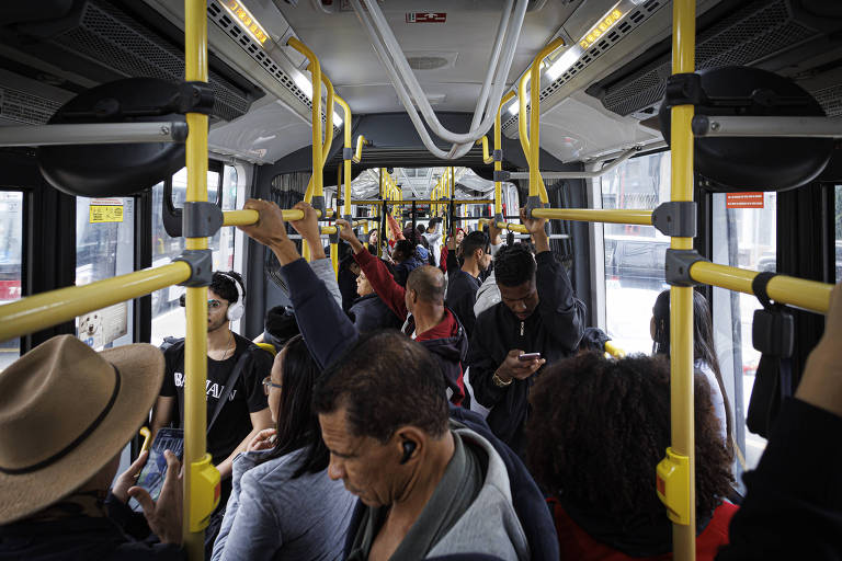 A imagem mostra o interior de um ônibus urbano lotado, com várias pessoas em pé e sentadas. As barras de apoio são amarelas e estão visíveis ao longo do ônibus. Algumas pessoas estão segurando seus celulares, enquanto outras conversam. A iluminação é natural, entrando pelas janelas do ônibus.