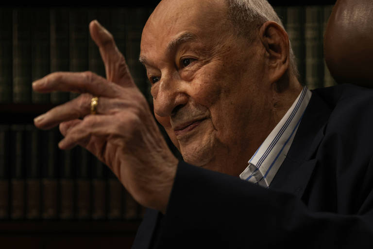 Um homem idoso está sentado， gesticulando com a mão direita levantada e o dedo indicador apontando para cima. Ele usa um paletó escuro e uma camisa listrada. Ao fundo， há prateleiras com livros. O ambiente é bem iluminado， destacando a expressão facial do homem， que parece estar em um momento de reflexão ou conversa.