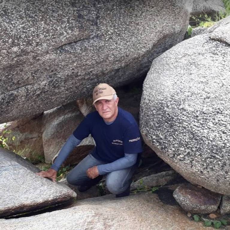 Uma pessoa está agachada entre grandes rochas. A pessoa usa uma camiseta azul e um boné. O ambiente é natural， com vegetação ao fundo e várias pedras ao redor.