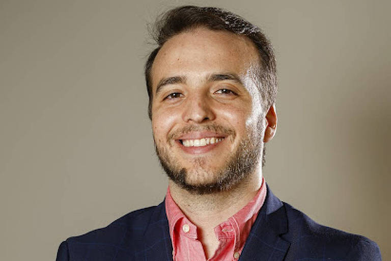 Homem branco， de cabelo castanho liso e barba rala， com os braços cruzados， vestido de paletó azul marinho e camisa de linho avermelhada