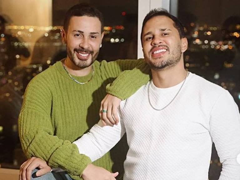 A imagem mostra dois homens sorrindo， um ao lado do outro， em um ambiente interno com uma vista da cidade ao fundo. O homem à esquerda está usando um suéter verde e o homem à direita está vestindo um suéter branco. Ambos estão com os braços apoiados um no outro e parecem estar se divertindo.