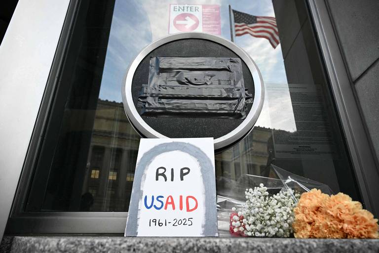 A imagem mostra uma janela com um círculo preto no centro， simbolizando um caixão. Em frente à janela， há um cartaz que diz 039;RIP USAID 1961-2025039;， desenhado com letras coloridas. Ao lado do cartaz， há flores， incluindo flores brancas e laranjas. Ao fundo， é visível uma bandeira americana e um sinal de entrada.