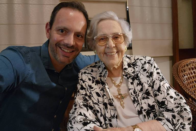 A imagem mostra um homem sorridente ao lado de uma mulher idosa， também sorridente. O homem está usando uma camisa azul e a mulher está vestindo um casaco com estampas florais em preto e branco. Eles estão sentados em um ambiente interno， com cortinas ao fundo.