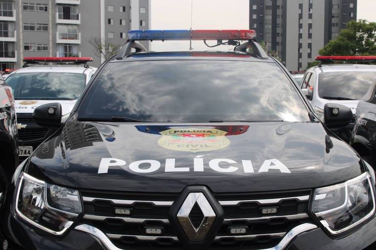 A imagem mostra uma viatura da polícia， modelo Renault， com a palavra 039;POLÍCIA039; em destaque na parte frontal. O veículo é preto e possui luzes de emergência vermelha e azul no teto. Ao fundo， é possível ver outros veículos e prédios.