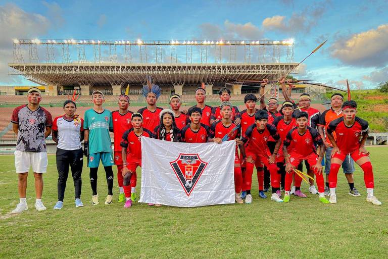 Uma equipe de futebol posando em um campo. Os jogadores estão vestidos com uniformes vermelhos e pretos， e alguns usam adereços como penas e pinturas faciais. No centro， há uma bandeira com o logo do GKFC. Ao fundo， um estádio com arquibancadas e céu parcialmente nublado.