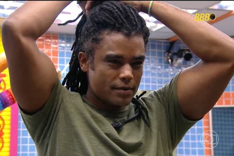 Um homem com dreadlocks está segurando o cabelo para cima, em um ambiente colorido. Ele usa uma camiseta verde e parece concentrado. Ao fundo, há paredes em tons de azul e amarelo, além de objetos coloridos que parecem ser parte de um jogo ou decoração.