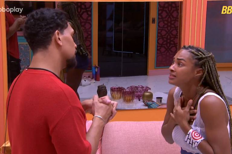 A imagem mostra duas pessoas em uma conversa intensa em um ambiente interno. Um homem, de costas, usa uma camiseta vermelha e está com as mãos juntas, enquanto uma mulher, de frente, usa uma blusa branca e parece estar emocionada, com as mãos no peito. Ao fundo, há uma decoração colorida e alguns objetos sobre uma mesa.