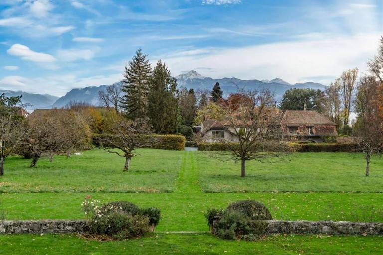 A imagem mostra uma paisagem de campo com um grande gramado verde， árvores dispersas e uma casa ao fundo. As montanhas são visíveis ao longe sob um céu parcialmente nublado. O ambiente parece tranquilo e natural， com algumas flores e arbustos na parte inferior da imagem.