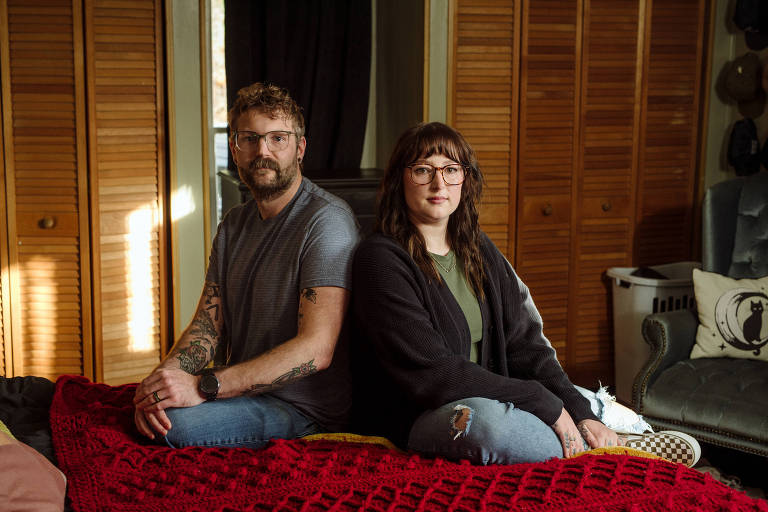 A imagem mostra um casal sentado em uma cama coberta com um edredom vermelho. Eles estão posicionados de costas um para o outro， com expressões neutras. O homem à esquerda tem cabelo curto e barba， vestindo uma camiseta cinza e jeans. A mulher à direita tem cabelo longo e escuro， usando uma blusa verde e um casaco preto. O ambiente é um quarto com paredes de madeira e móveis ao fundo.