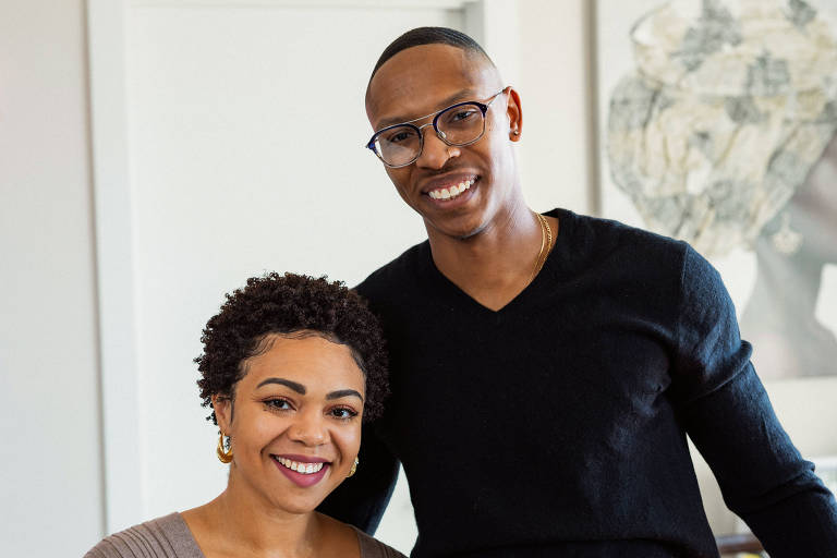 A imagem mostra um casal posando em um ambiente interno. A mulher está à esquerda， com cabelo curto e encaracolado， vestindo um suéter de cor clara. O homem está à direita， sorrindo， usando uma camiseta preta e jeans. Ao fundo， há uma parede clara e uma obra de arte decorativa.