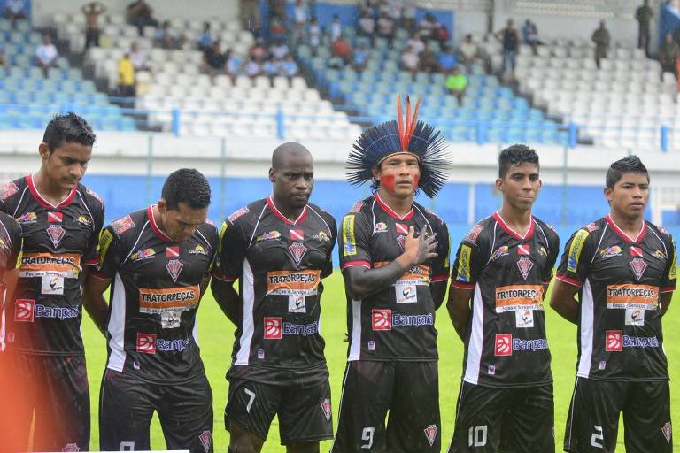 Um grupo de seis jogadores de futebol está alinhado em um campo， todos vestindo uniformes pretos com detalhes em vermelho e amarelo. O jogador no centro usa um cocar colorido e tem pintura facial. Eles estão em uma posição respeitosa， com as mãos sobre o coração， possivelmente durante a execução do hino nacional. Ao fundo， há uma arquibancada com algumas pessoas assistindo ao evento sob chuva.