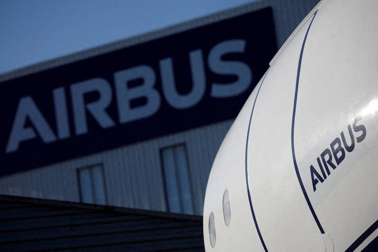 A imagem mostra uma parte de um edifício com o logotipo da Airbus em destaque. O logotipo é grande e está em letras brancas sobre um fundo azul escuro. Na frente， há uma parte de um objeto， possivelmente uma fuselagem de aeronave， com o nome 039;AIRBUS039; visível.