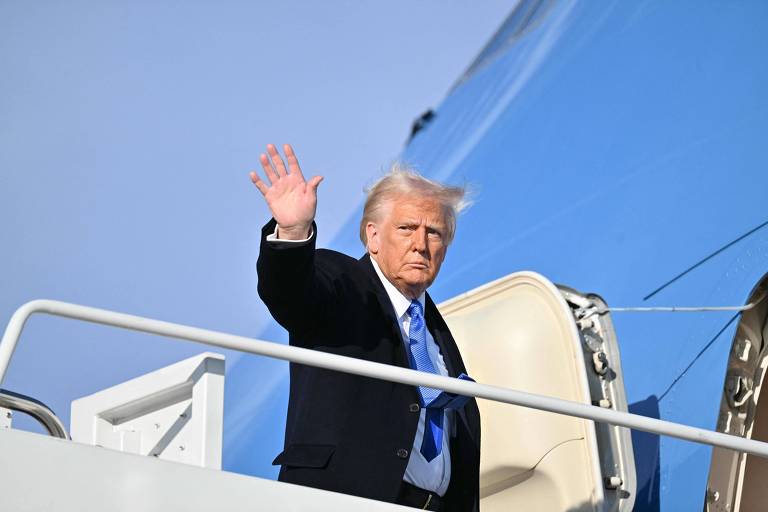 Um homem branco está acenando com a mão enquanto está na escada de um avião. Ele usa um terno escuro e uma gravata azul. O fundo é um céu claro e o avião tem uma parte azul visível.