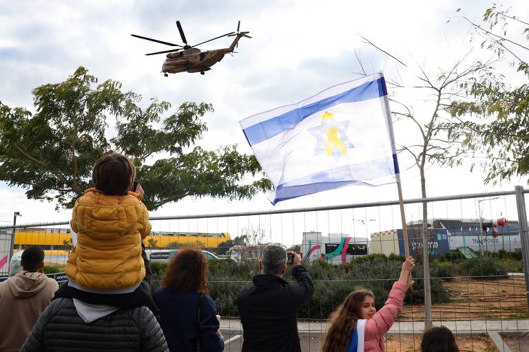 Um grupo de pessoas está assistindo a um helicóptero que sobrevoa a área. Uma criança está sentada nos ombros de um adulto, enquanto outras pessoas seguram uma bandeira. Ao fundo, há árvores e uma cerca. O céu está nublado.