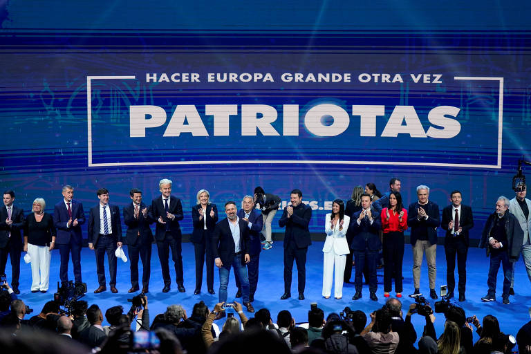 A imagem mostra um grupo de pessoas em um palco durante um evento político. Ao fundo， há um fundo com o texto 039;HACER EUROPA GRANDE OTRA VEZ039; e a palavra 039;PATRIOTAS039; em destaque. As pessoas estão aplaudindo e algumas estão vestidas formalmente， enquanto outras usam roupas mais casuais. O ambiente é iluminado e há câmeras registrando o momento.