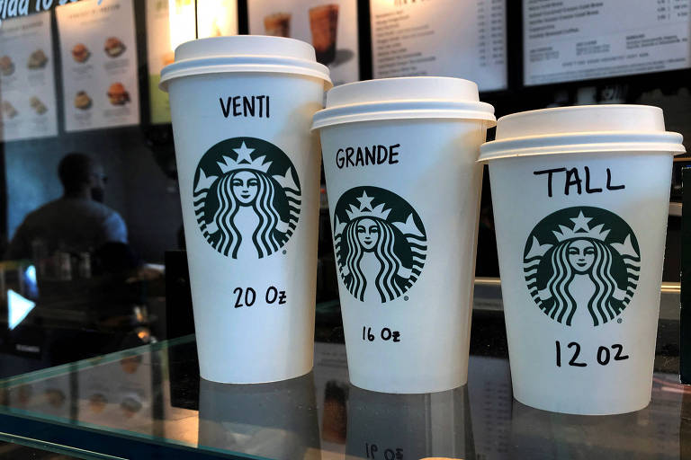 A imagem mostra três copos de café da Starbucks dispostos em uma superfície. O copo da esquerda é rotulado como 039;VENTI039; com a capacidade de 20 oz， o copo do meio é rotulado como 039;GRANDE039; com a capacidade de 16 oz， e o copo da direita é rotulado como 039;TALL039; com a capacidade de 12 oz. Todos os copos têm o logotipo da Starbucks impresso na frente.