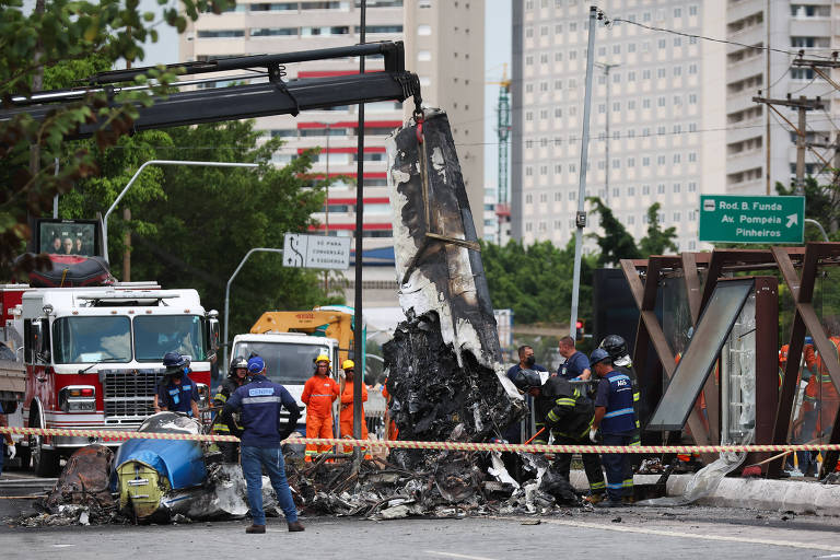 A imagem mostra uma cena de acidente em uma área urbana， com destroços de um avião ou estrutura danificada no centro. Há várias pessoas vestindo uniformes de emergência， incluindo bombeiros e equipes de resgate， trabalhando na cena. Veículos de emergência， como caminhões de bombeiros， estão presentes ao fundo， e edifícios altos são visíveis nas proximidades. A área está isolada com fita de segurança