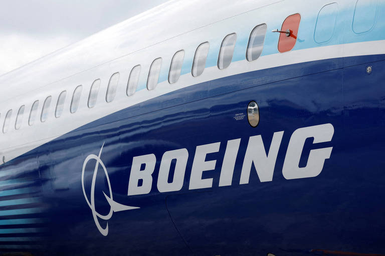 A imagem mostra a fuselagem de um avião da Boeing， destacando o logotipo da empresa em letras grandes e brancas. O fundo é de um azul escuro， e há detalhes em cinza e vermelho na estrutura do avião.