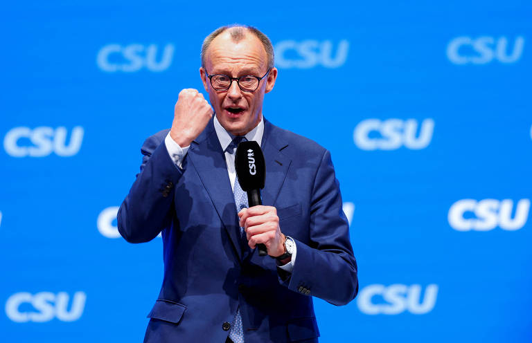 Um homem em um terno azul está em um palco， segurando um microfone e fazendo um gesto de punho cerrado. O fundo é azul com a sigla 039;CSU039; repetida em branco.