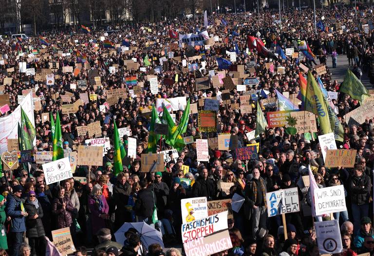 A imagem mostra uma grande multidão reunida em uma manifestação. As pessoas seguram cartazes e bandeiras， com uma variedade de mensagens visíveis. A cena é de um evento ao ar livre， com muitos participantes visíveis em um espaço amplo.