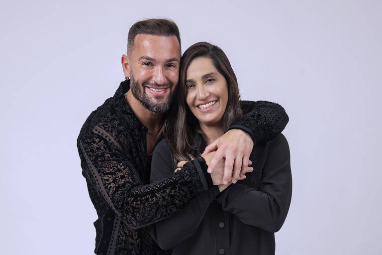 Um homem e uma mulher estão posando juntos em um fundo branco. O homem está usando uma camisa preta com detalhes em veludo e calças pretas, enquanto a mulher está vestindo uma blusa preta e uma saia curta preta. Eles estão sorrindo e o homem está abraçando a mulher, que está segurando suas mãos.