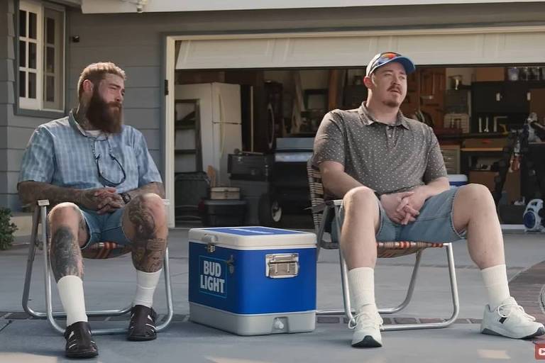 A imagem mostra dois homens sentados em cadeiras de praia em um ambiente externo. O homem à esquerda usa uma camisa de estampa e um boné azul com vermelho， enquanto o homem à direita tem uma barba e usa uma camisa listrada. Ambos parecem estar relaxando e observando algo fora do quadro.