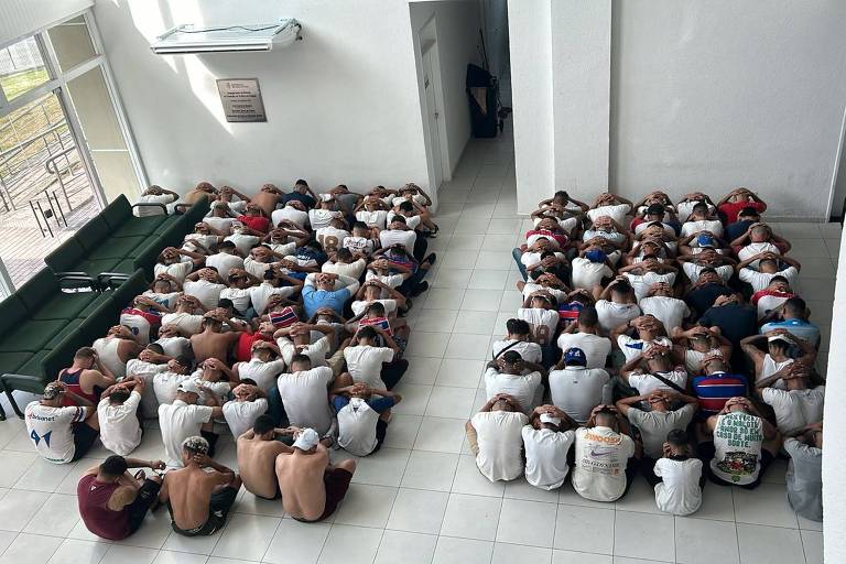 A imagem mostra um grande grupo de pessoas sentadas no chão， com as mãos na cabeça， em um ambiente interno. Elas estão organizadas em duas seções， uma à esquerda e outra à direita， com um sofá verde ao fundo. O ambiente é bem iluminado， com janelas grandes que permitem a entrada de luz natural. O piso é de cerâmica branca.