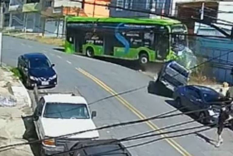 A imagem mostra uma rua com vários veículos estacionados e em movimento. Há um ônibus verde e branco， além de carros de diferentes cores， como preto e branco. O ambiente parece ser urbano， com edifícios e comércio ao fundo. A iluminação é clara， indicando que é dia.